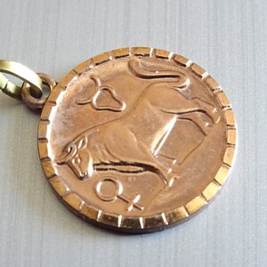 Pode incluir: Um pingente de metal dourado com uma representação detalhada de um touro, símbolo do signo do zodíaco Touro. O pingente tem uma borda texturizada e um pequeno símbolo feminino na parte inferior.