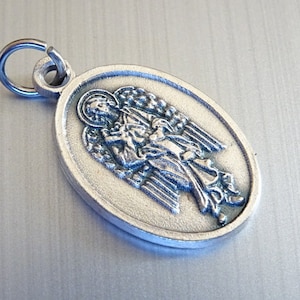 May include: Silver oval pendant featuring a detailed depiction of an angel with wings outstretched. The pendant has a loop at the top for attaching to a chain.