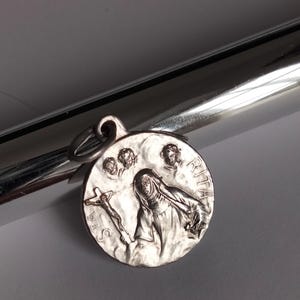 May include: Silver pendant featuring a relief image of a woman in a veil, holding a cross. The text "STA RITA" is inscribed around the edge of the pendant.