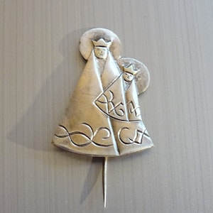 May include: Silver brooch featuring a depiction of the Virgin Mary and baby Jesus. The brooch is detailed with intricate designs and a pin on the back.
