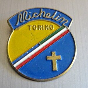 Könnte beinhalten: Eine blaue und gelbe runde Anstecknadel mit dem Wort "Michelin" in Kursivschrift und dem Wort "Torino" darunter. Die Anstecknadel hat einen roten, weißen und grünen Streifen darüber und ein goldenes Kreuz in der Mitte.