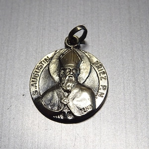 Op de afbeelding: Zilverkleurige metalen hanger met een reliëfbeeld van Sint Augustinus, een katholieke heilige, die een miter draagt en een boek vasthoudt. De hanger is gegraveerd met "S. AUGUSTIN" en "PRIEZ P.N."