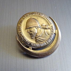 May include: A gold-colored metal pin with a crescent moon design. The pin features a profile of a soldier wearing a helmet and carrying a rifle. The text around the soldier reads "Tout Ancien Combattant Aide et Secours a Son Camarade  Poilu".
