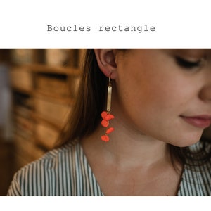 Peut inclure: Une paire de boucles d'oreilles pendantes avec une barre dorée et plusieurs breloques en forme de fleurs orange vif. Les boucles d'oreilles sont portées par une personne aux cheveux longs et bruns.