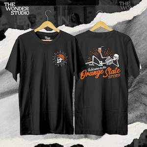 Puede incluir: Camiseta negra con un gráfico de calavera en la parte delantera y un gráfico de esqueleto en la parte trasera. El esqueleto está tumbado con una bebida en la mano. El texto "Welcome to the Orange State" está impreso debajo del esqueleto. El texto "SUN SCREAM" está impreso en la parte inferior izquierda de la imagen. El texto "BAD TO THE BONE FRIDAY 08.12" está impreso en la parte inferior derecha de la imagen.
