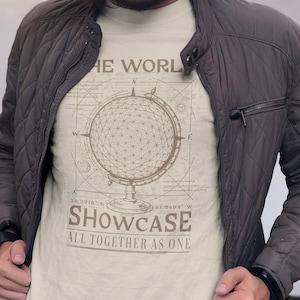 Könnte beinhalten: Beiges T-Shirt mit einem Globus-Design und dem Text "THE WORLD SHOWCASE ALL TOGETHER AS ONE". Das Shirt wird unter einer dunkelbraunen, gesteppten Jacke getragen. Das Globus-Design enthält eine Windrose und Koordinaten.