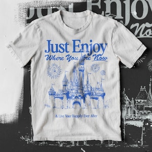 May include: A white t-shirt with a blue graphic of a castle and fireworks. The text "Just Enjoy Where You Are Now & Live Your Happily Ever After" is printed on the shirt.