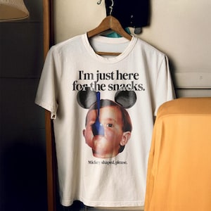 Puede incluir: Camiseta blanca con un gr&aacute;fico de un ni&ntilde;o con orejas de Mickey Mouse comiendo con una cuchara azul. El texto en la camiseta dice "I'm just here for the snacks. Mickey shaped, please."