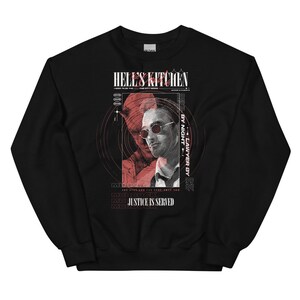 Könnte beinhalten: Schwarzes Sweatshirt mit einem grafischen Design. Das Design enthält den Text "HELL'S KITCHEN" und "JUSTICE IS SERVED", ein Porträt eines Mannes mit Sonnenbrille und eine rote Silhouette. Das Sweatshirt hat einen Rundhalsausschnitt.