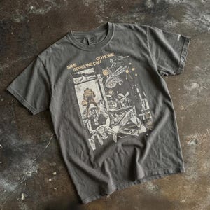 May include: Dark grey t-shirt with a graphic design featuring the text "SAVE STARS, WE CAN GO HOME." The design includes illustrations of figures and machinery. The shirt is made of a soft material and has a crew neck.