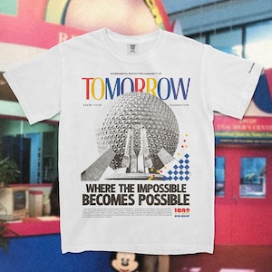 May include: White t-shirt with a graphic print of the Spaceship Earth attraction at Epcot. The graphic is in black and white with red, blue, and yellow accents. The text on the shirt reads "EXPERIMENTAL PROTOTYPE COMMUNITY OF TOMORROW" and "WHERE THE IMPOSSIBLE BECOMES POSSIBLE".