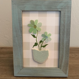 May include: A framed artwork featuring a light blue wooden frame. The artwork inside has a checkered background with a light green flower design. The flowers are in a light green pot.