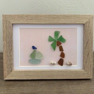 May include: A framed artwork featuring a beach scene. A sailboat with a blue flag and a palm tree are crafted from sea glass. Two seashells are at the bottom. The frame is light brown.