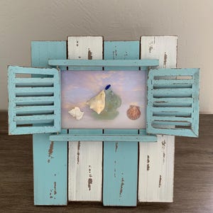 May include: A decorative wall hanging featuring a distressed light blue and white wooden frame designed like a window with open shutters. The center displays a sunset sky print adorned with three seashells and a small piece of blue sea glass.