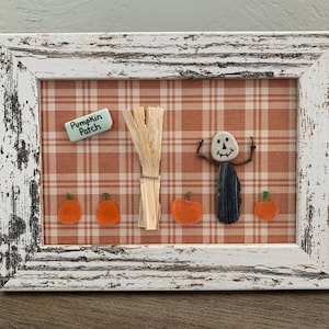 May include: A framed autumn-themed artwork with a distressed white wooden frame. The artwork features a plaid background, a straw bundle, a stone scarecrow, orange pumpkins, and a sign that reads "Pumpkin Patch".