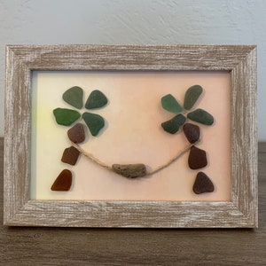 May include: A framed artwork featuring two palm trees made from sea glass and a hammock made from driftwood. The background is a gradient of pink and yellow.
