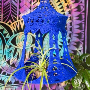 May include: A blue crocheted hanging planter with a plant inside. The planter is hanging in front of a colorful mandala tapestry.