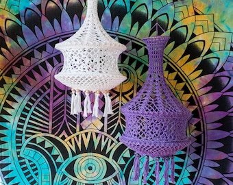 May include: Two crocheted lampshades hanging against a colorful mandala background. The lampshades are made of white and purple yarn and have a delicate, intricate design.