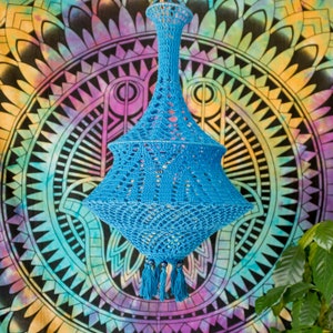 May include: A blue crocheted lampshade with tassels hanging from the bottom. The lampshade is hanging from the ceiling against a colorful mandala background.