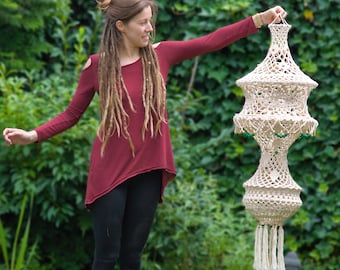 May include: A cream macrame hanging lantern with a tiered design and a tassel fringe. The lantern is held up against a backdrop of green foliage. The lantern has a decorative, open weave pattern and is suspended from a loop at the top.