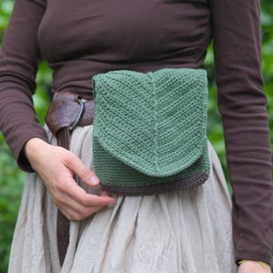 Puede incluir: Un cinturón de ganchillo verde con una solapa en forma de hoja, usado alrededor de la cintura. El bolso es de color verde liso con un ribete marrón. La persona lleva una camisa marrón de manga larga y una falda beige.