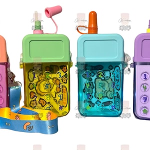 May include: Four colorful, clear plastic water bottles shaped like old-fashioned cell phones with a straw and a carrying strap. The bottles have different designs on them, including numbers, cartoon animals, and other fun graphics.