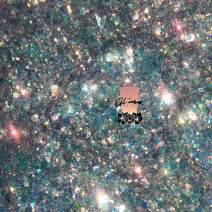 May include: A close-up of iridescent blue, green, and silver glitter. The glitter is scattered on a white surface. The text "Glamour Designs" is visible in the image.