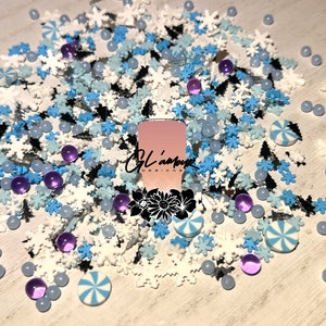 May include: A variety of winter-themed sprinkles in white, blue, and black. The sprinkles include snowflakes, candy canes, and small beads. There is a pink square with the text "Glamour Creations" in black.