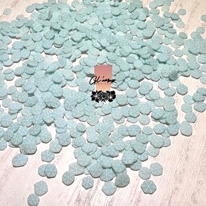 May include: A large pile of light blue snowflake sprinkles. The sprinkles are scattered on a white surface.