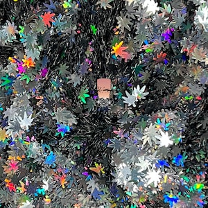 May include: A close-up of iridescent holographic glitter in the shape of marijuana leaves. The glitter is scattered on a black surface.