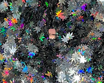 May include: A close-up of iridescent holographic glitter in the shape of marijuana leaves. The glitter is scattered on a black surface.