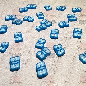 May include: A collection of blue butterfly-shaped resin embellishments. The embellishments are scattered on a light wood surface.