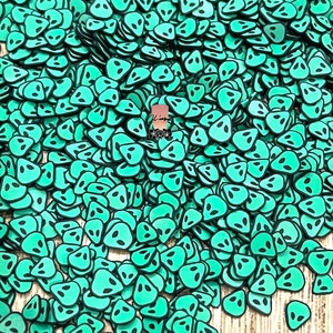 May include: A large pile of green alien face shaped resin flatbacks. The alien faces have black eyes and are scattered on a wood surface.