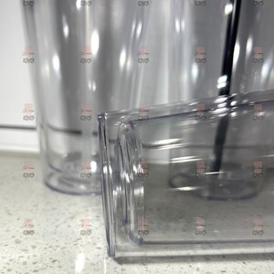 Puede incluir: Vasos de pl&aacute;stico transparente con base rectangular y cuerpo cil&iacute;ndrico. Los vasos son transparentes, lo que permite ver el contenido. Se ve una pajita negra en uno de los vasos. Los vasos est&aacute;n sobre una superficie blanca.