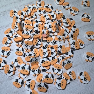 May include: A collection of small, cow-shaped embellishments. Each cow has a white face with black spots, tan ears, and a tan muzzle. The embellishments are scattered on a light-colored surface.