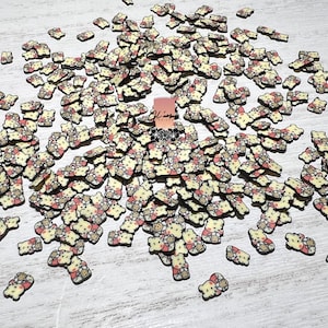 May include: A large quantity of small, yellow and pink resin charms shaped like cartoon bears. The charms have black outlines and are scattered on a light wood surface.