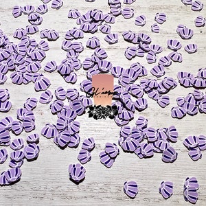May include: A large quantity of purple and white seashell shaped resin embellishments. The seashells are scattered on a white wooden surface.