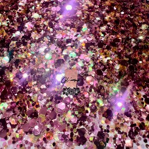 May include: A close-up of a pile of iridescent glitter in shades of pink, purple, and gold. The glitter is scattered on a white surface.