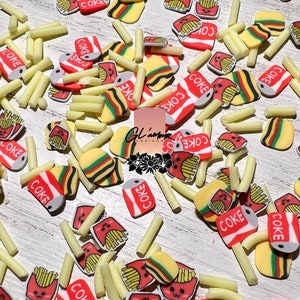 May include: A collection of miniature food-themed charms scattered across a light-colored surface. The charms include yellow french fries, red and white soda cans with "COKE" text, small hamburgers, and smiling french fry boxes.