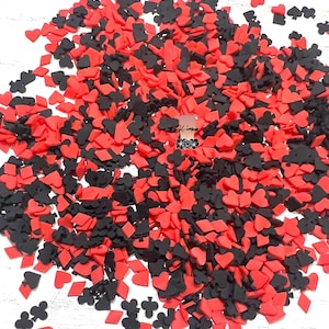 May include: A pile of red and black card suit sprinkles. The sprinkles are shaped like hearts, diamonds, clubs, and spades.