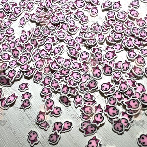 May include: A large quantity of pink and white skull-shaped resin embellishments with a small pink bow on each skull. The embellishments are scattered on a white surface.