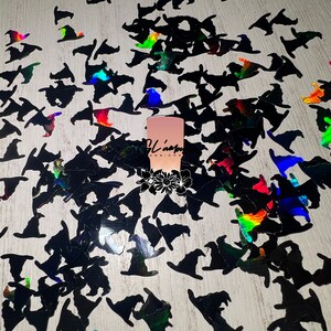 May include: A large quantity of black witch hat shaped confetti with iridescent rainbow glitter. The confetti is scattered on a light wood surface.