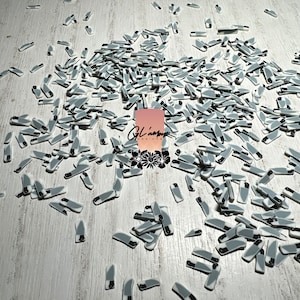 May include: A large quantity of small, gray and white knife-shaped charms. The charms are scattered on a light wood surface.