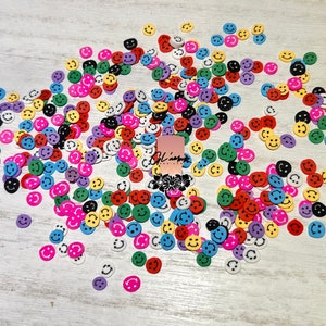 May include: A large group of colorful smiley face buttons scattered on a white wooden surface. The buttons are various shades of red, yellow, green, blue, purple, and black.