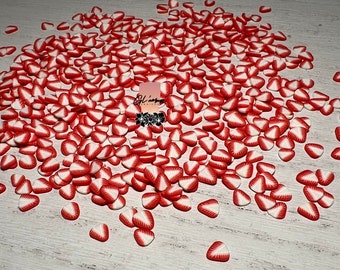 May include: A large pile of red and white strawberry slice shaped sprinkles. The sprinkles are scattered on a white surface.
