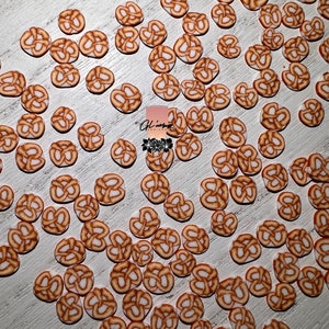 May include: A large collection of brown and white pretzel-shaped resin embellishments. The embellishments are scattered on a white wooden surface.