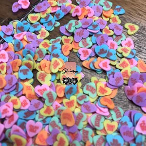 May include: A large pile of colorful heart-shaped candies with various messages printed on them, including "XOXO", "So Cute", "Be Mine", "Hug Me", and "I Love You".
