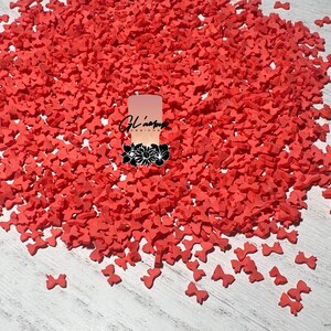 May include: A large pile of small, coral-colored bow-shaped sprinkles. The sprinkles are scattered across a light-colored wooden surface. A logo is visible in the center of the image.