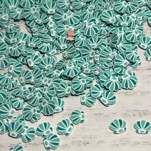 May include: A large pile of teal and white seashell shaped resin embellishments. The embellishments are scattered on a white wooden surface.
