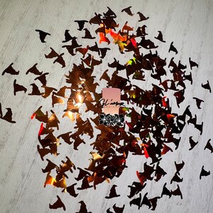 May include: A scattering of iridescent black witch hat shaped confetti on a white surface. The confetti is made of a holographic material that reflects light in a rainbow of colors.